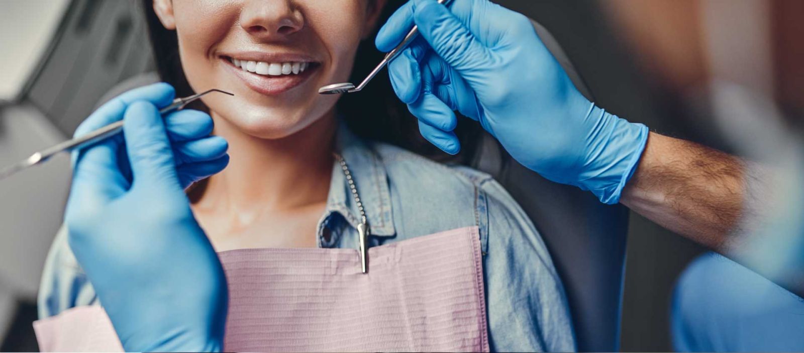 A dentist wearing blue gloves is holding tools to check the teeth of a woman with a nice smile.