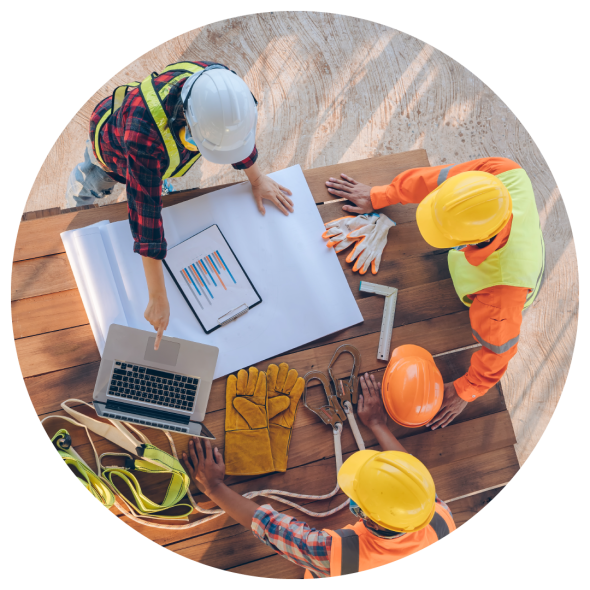 Construction workers wearing hard hats are gathered around a wooden table to plan a project using printed documents and a laptop computer.