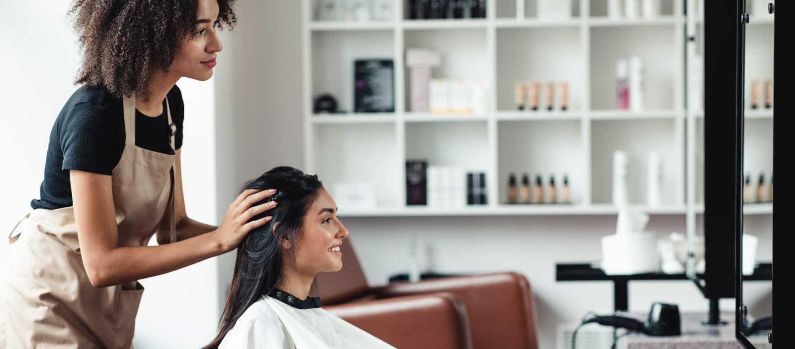 A hairdresser consults with her client while she is seated and wearing a white smock in a well lit salon.