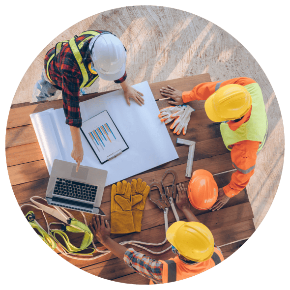 Construction workers wearing hard hats are gathered around a wooden table to plan a project using printed documents and a laptop computer.